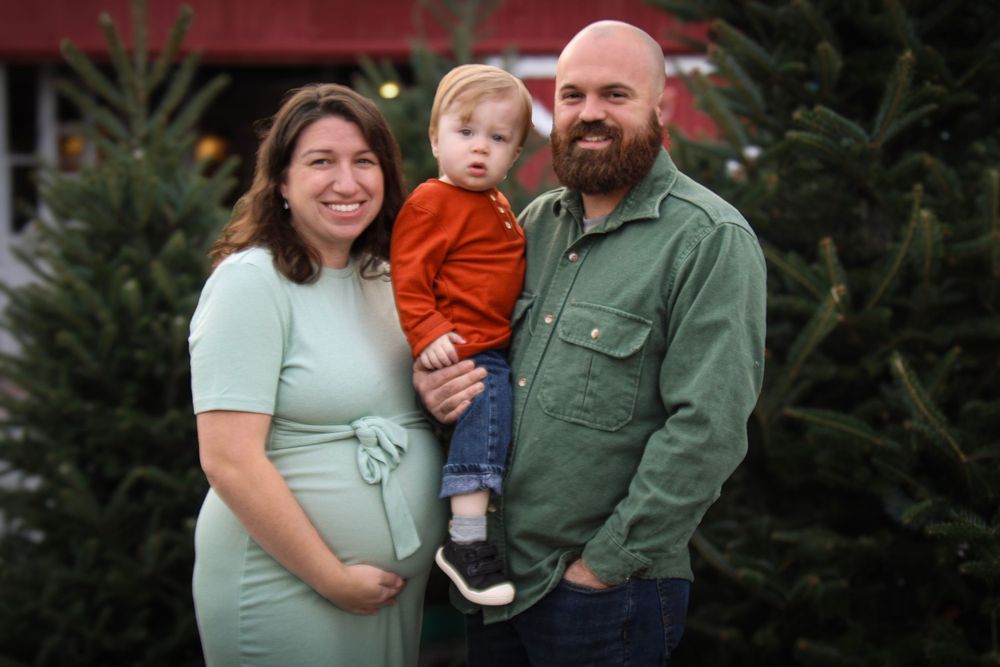 Family of three poses for photo at Christmas tree farm with red barn backdrop.