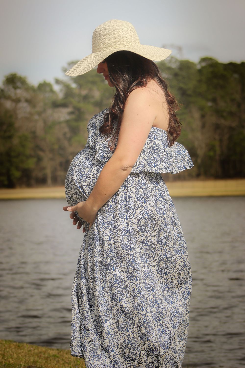 A figure in a flowing blue floral dress and sun hat stands near a serene lake on a sunny afternoon.