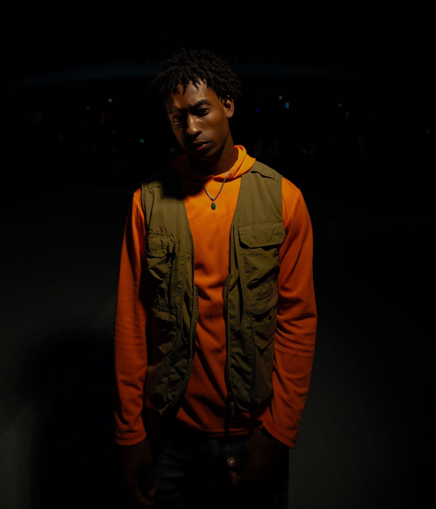 Dramatic portrait of someone wearing an orange sweater and olive utility vest in low-key lighting.