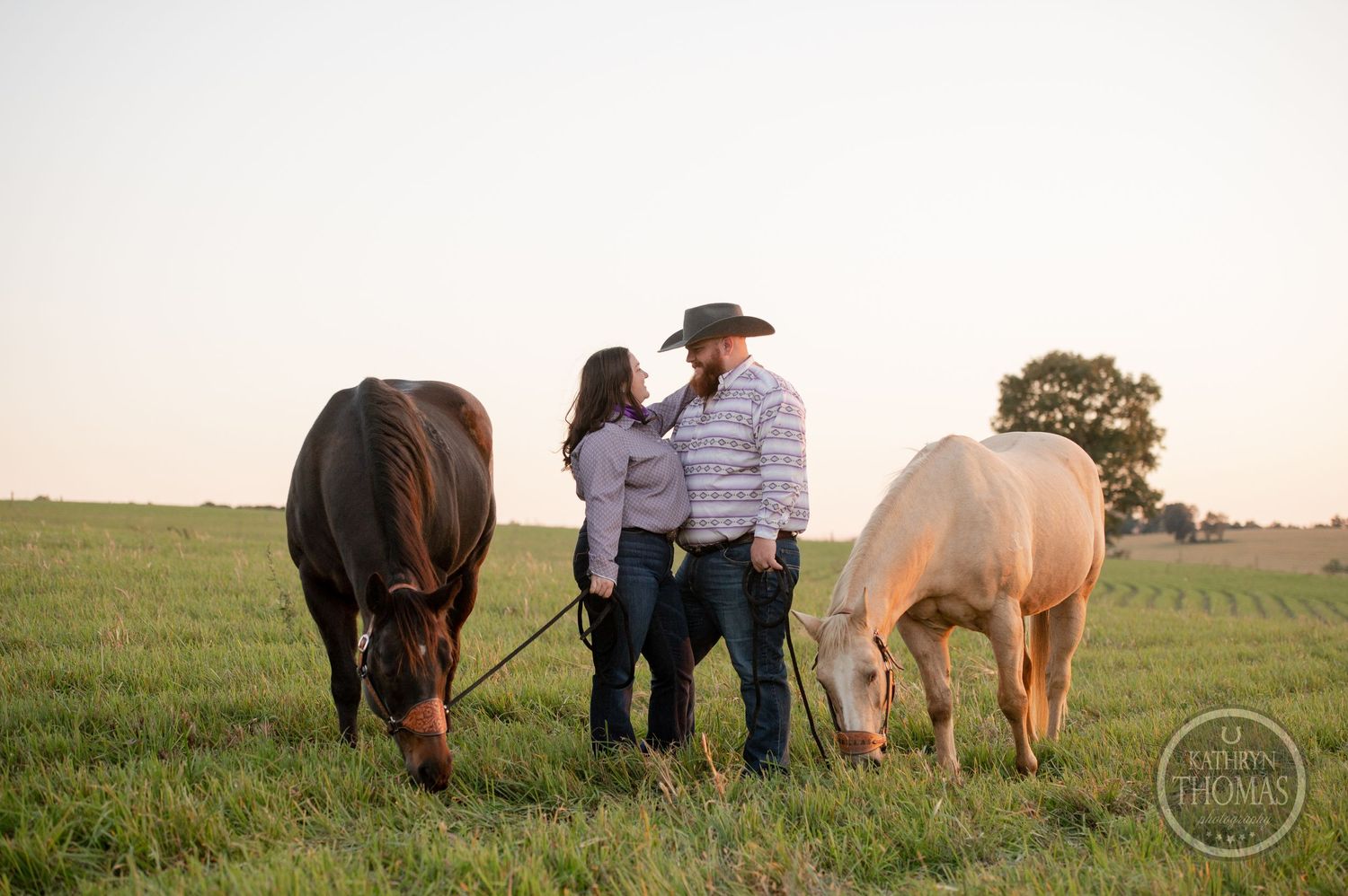 A western couple standing close together with their horses grazing in a peaceful field at sunset.