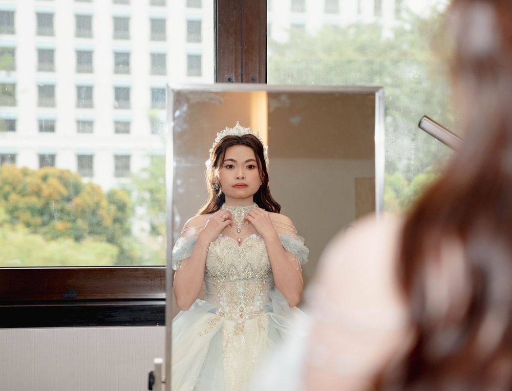 A bride in an elegant white lace gown adjusts her hair while standing by a window with city buildings visible outside.