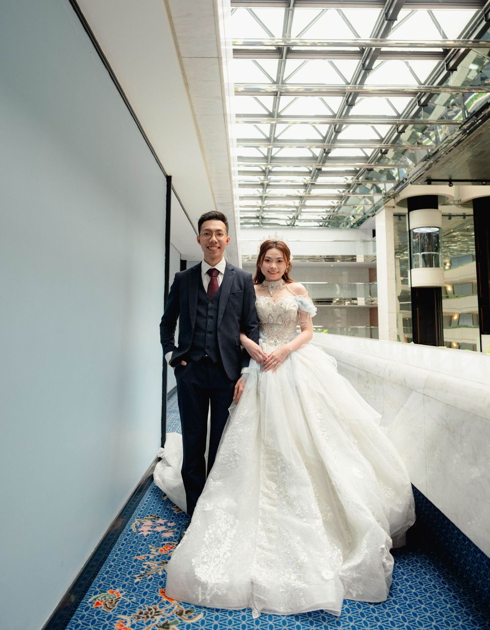 A couple poses in formal wedding attire on blue carpeted floor under a modern glass ceiling in an elegant indoor space.
