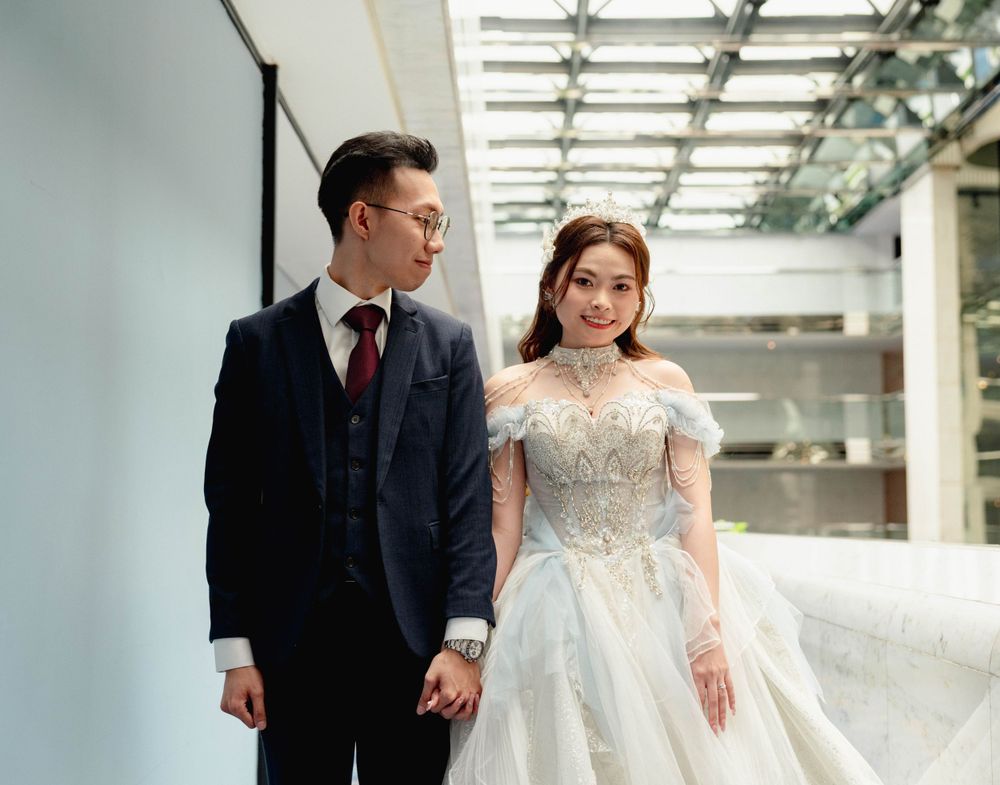 A couple in formal wedding attire holding hands in a modern building with glass ceiling and natural light.