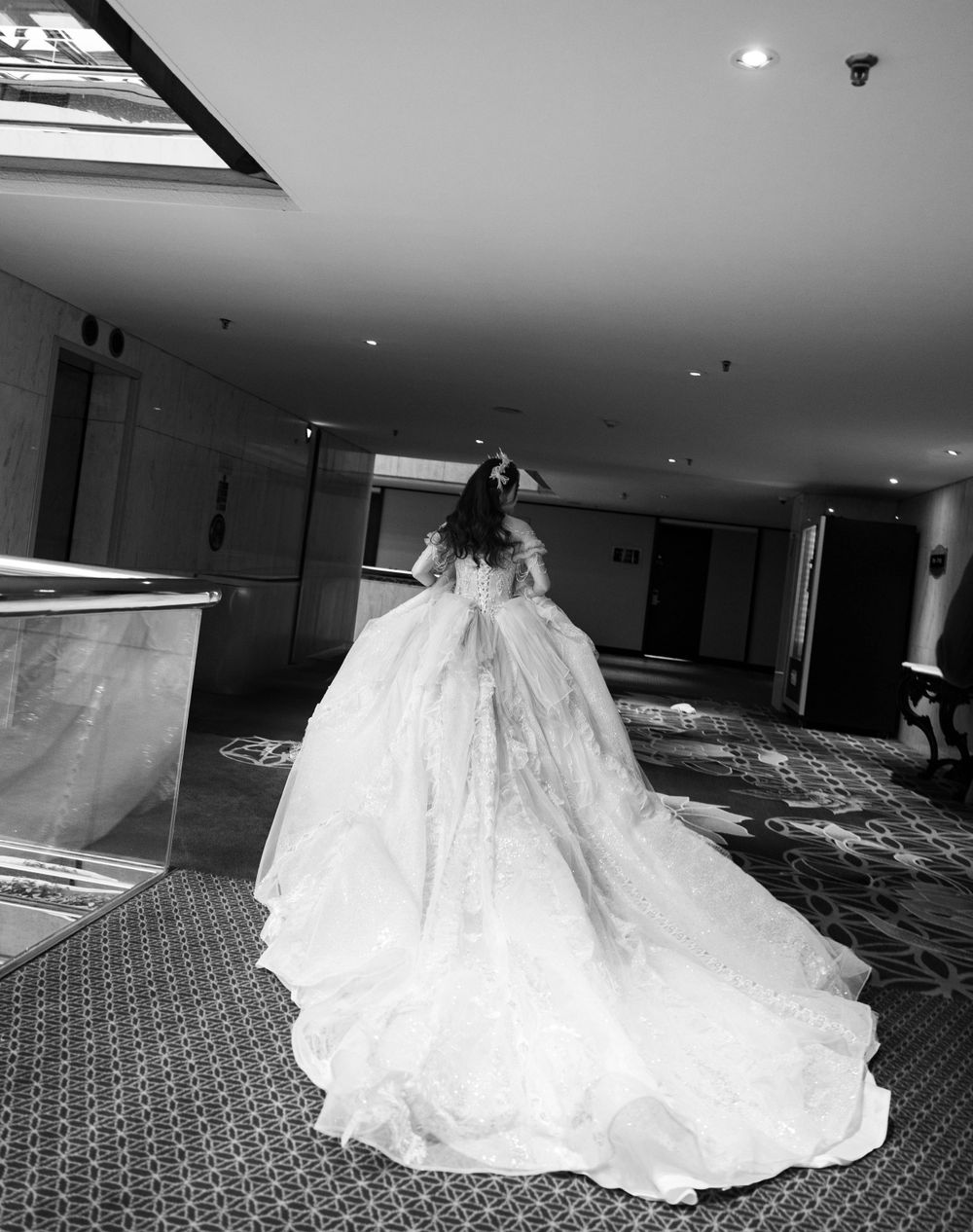 A dramatic full-length wedding dress with a voluminous skirt and long train captured in black and white in a hotel hallway.