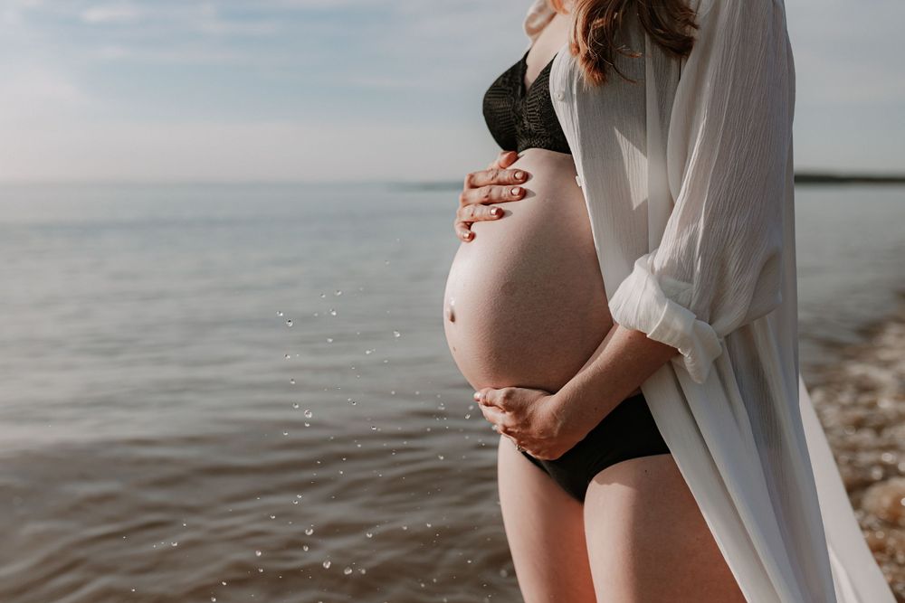 Pregnant woman in black bikini and white open shirt holding baby bump beside calm ocean.