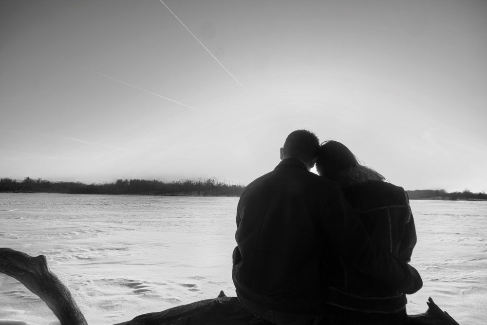 A couple sits close together in silhouette against a calm lake at dusk, creating a romantic black and white scene.