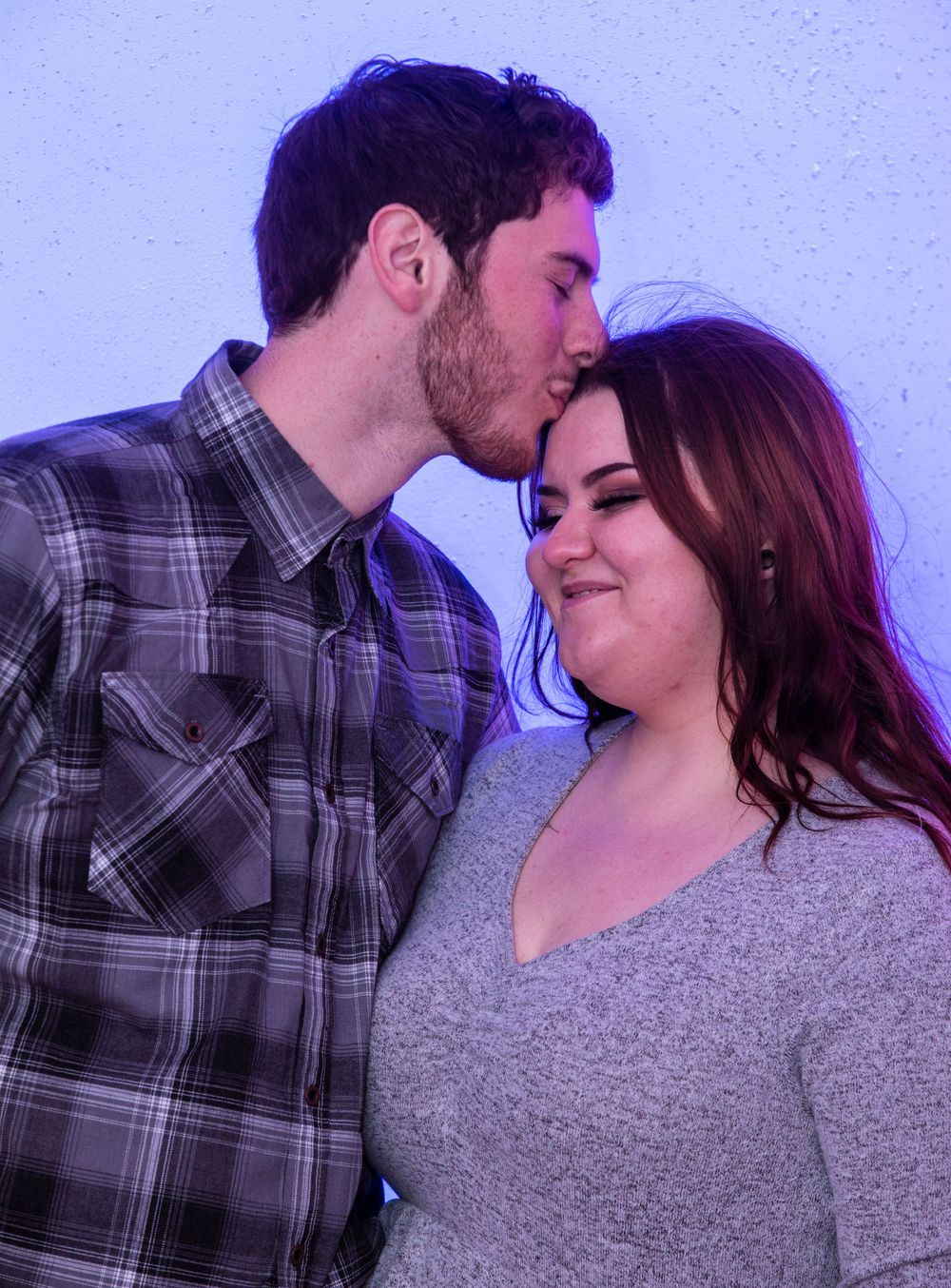 Two people share an intimate moment in purple lighting, with one wearing a plaid shirt and the other a gray sweater.