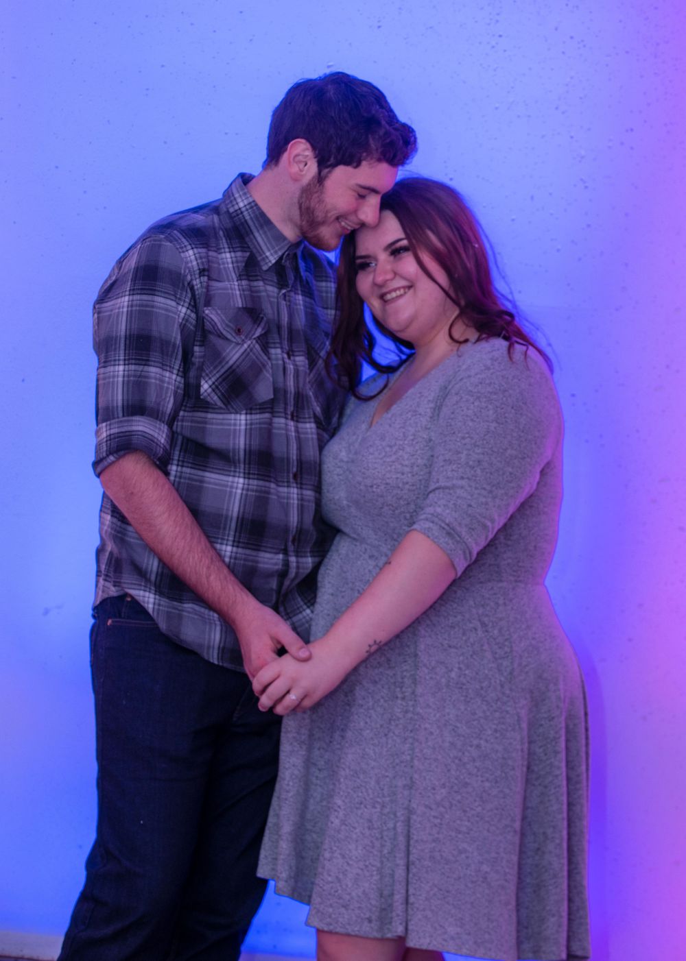 A couple shares an intimate moment against a purple-lit backdrop, with one wearing plaid and the other in a gray dress.