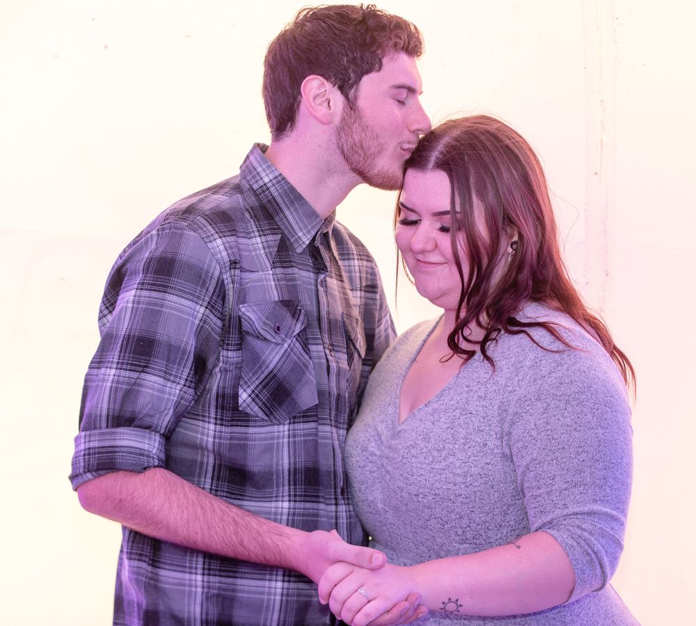 A tender moment between a couple where he wears a plaid shirt and she wears a gray dress against a light background.