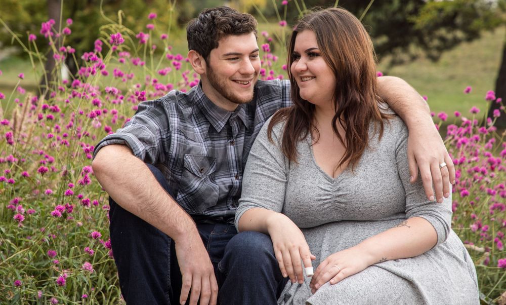 A couple sits together outdoors smiling on a sunny day with pink flowers blooming in the background.