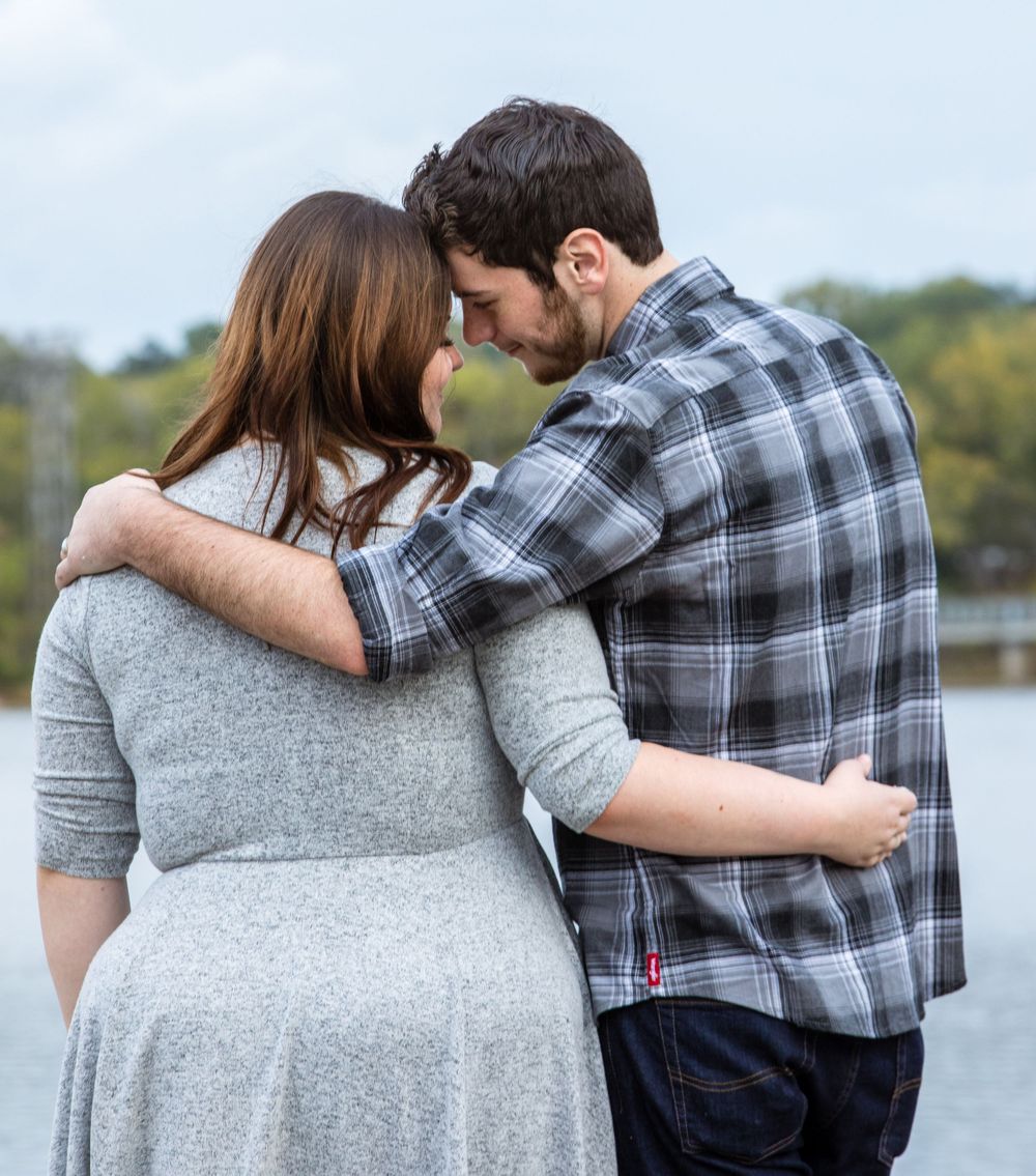 A couple in casual wear embraces while standing by a lake, with the person in plaid holding their partner in a gray dress.