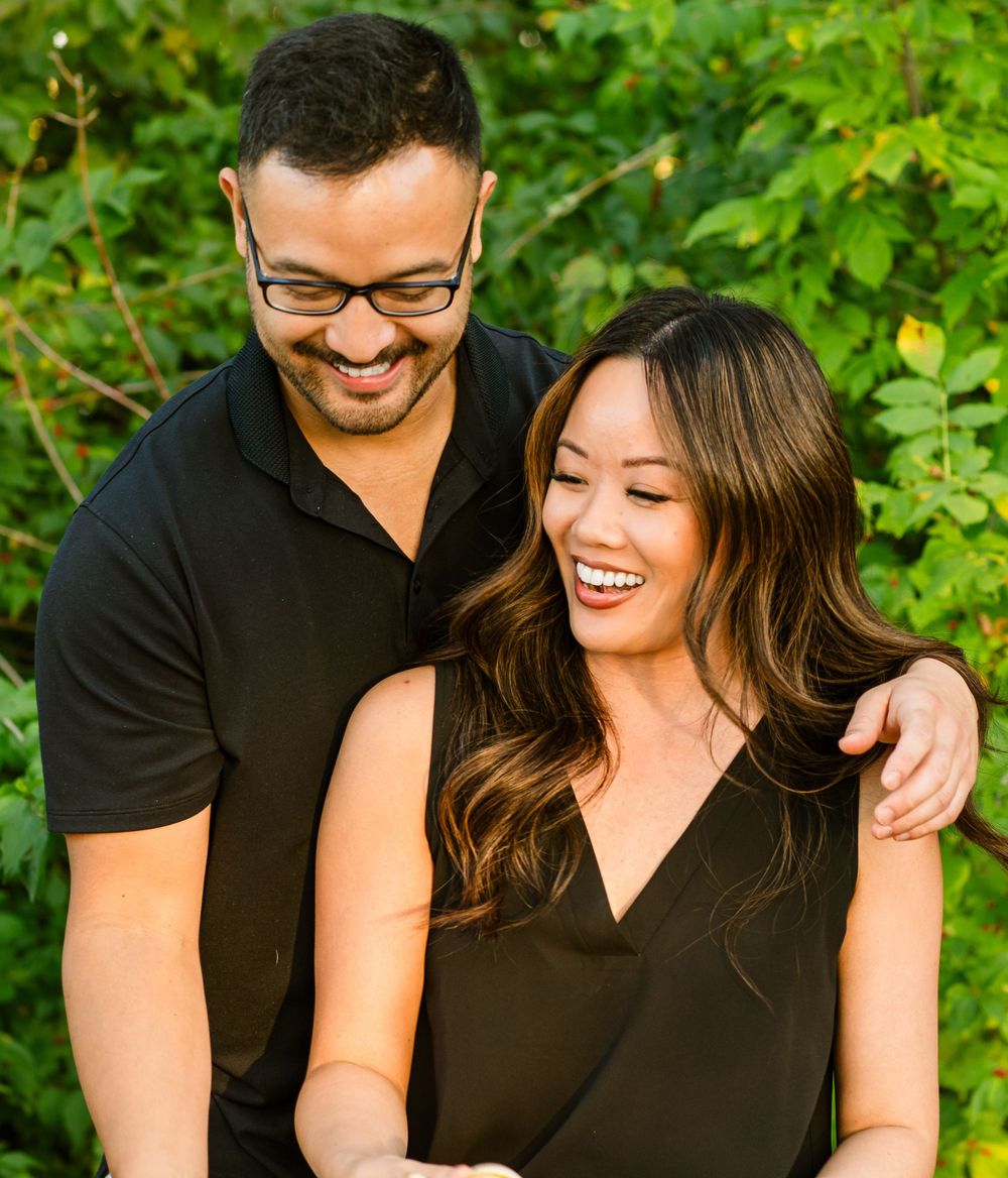 A couple wearing black outfits share a joyful candid moment together against green foliage in the background.