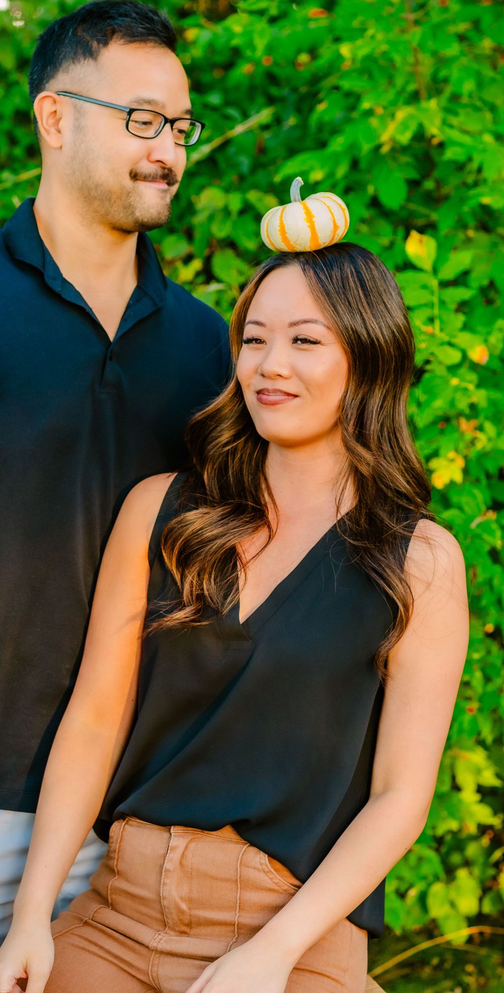 A couple in black shirts pose for a fall photo with a small decorative pumpkin balanced on her head against green foliage.