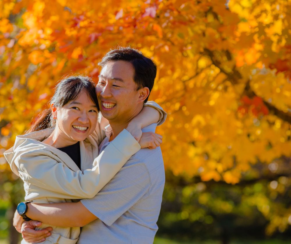 A couple embraces outdoors with bright orange autumn foliage in the background during golden hour.