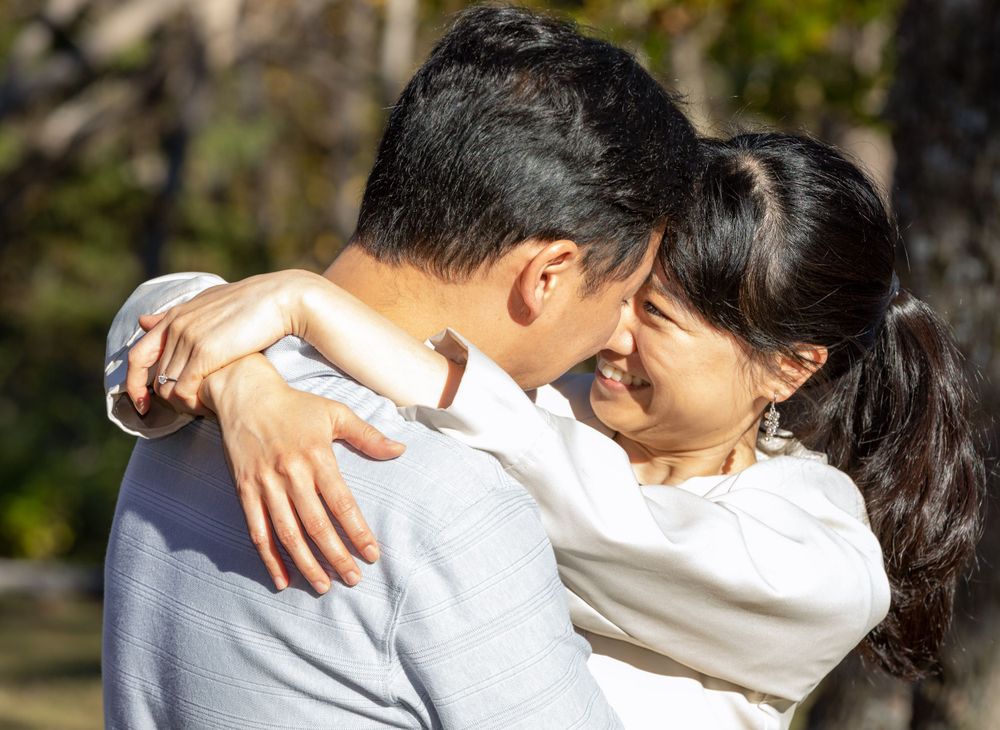 A couple embraces warmly in sunlit outdoor setting wearing white sweaters.
