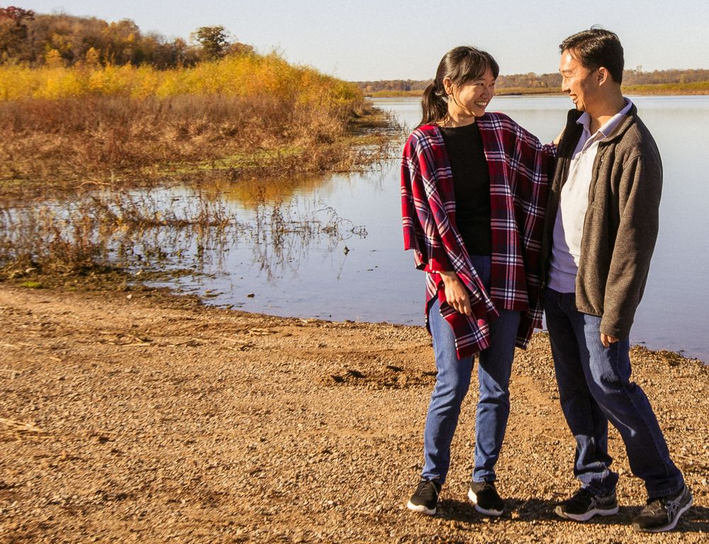 Two people enjoy an autumn walk along a serene lake shoreline with golden grasses and trees in the background.