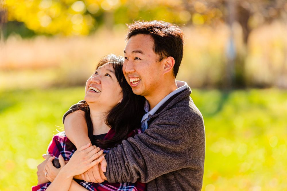 A couple shares a joyful embrace while laughing together in a sunlit autumn setting with yellow foliage in background.
