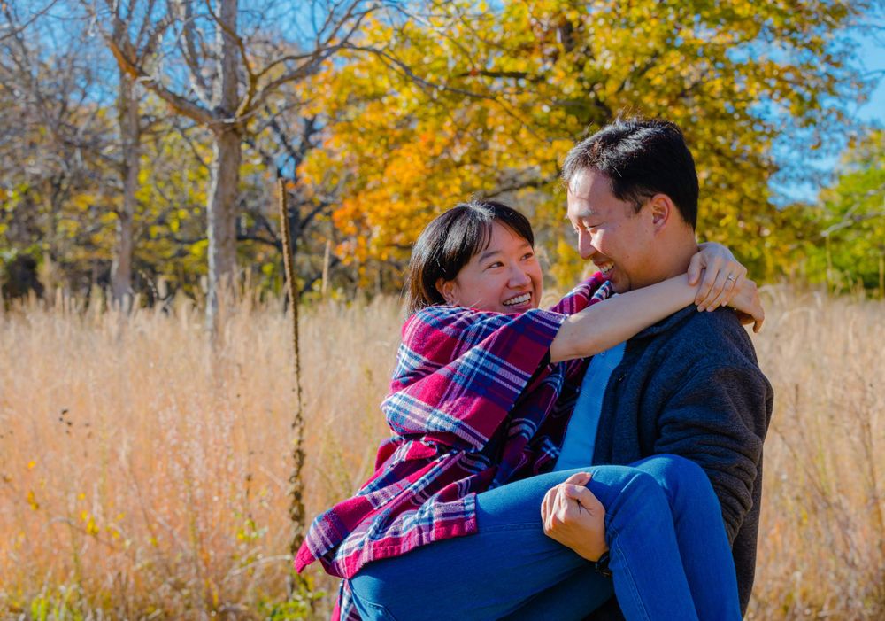 A romantic couple embraces playfully in a field with golden fall colors and autumn trees behind them.