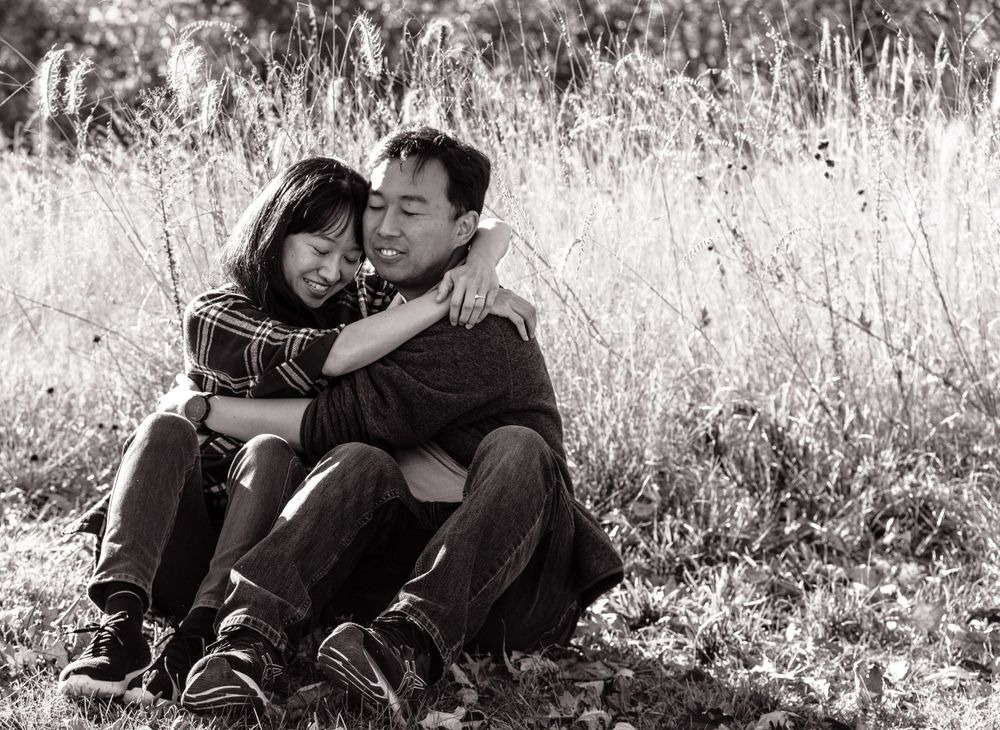 A black and white photo of a couple sitting in tall grass embracing and laughing.