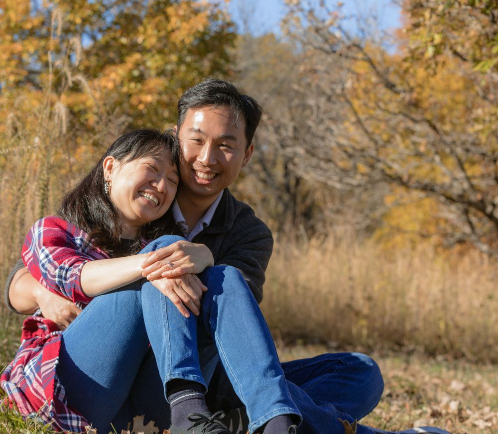 A couple sits together outdoors enjoying an autumn day with golden leaves and trees in the background.