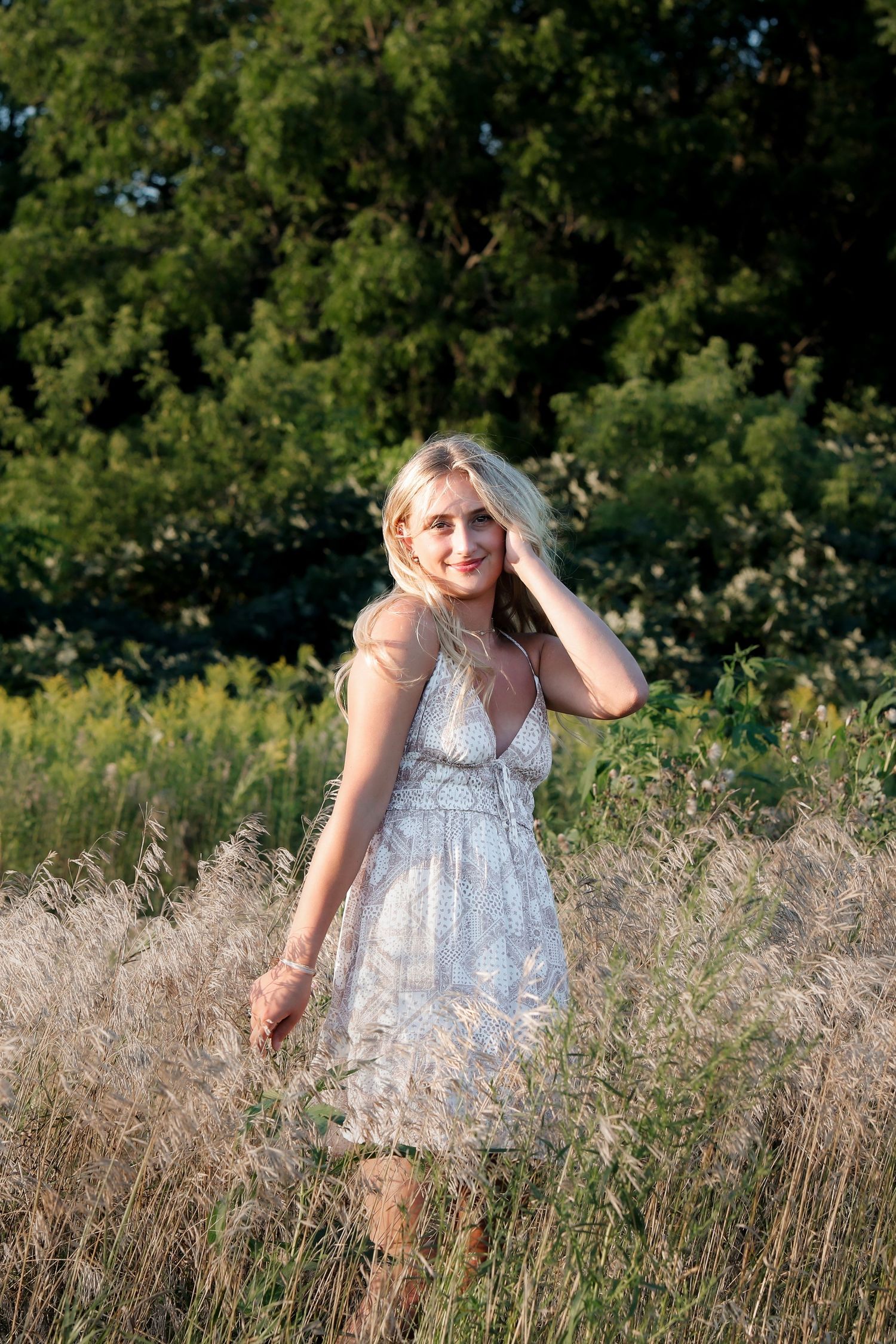 Blonde woman in white lace dress standing in golden field with green trees in background.