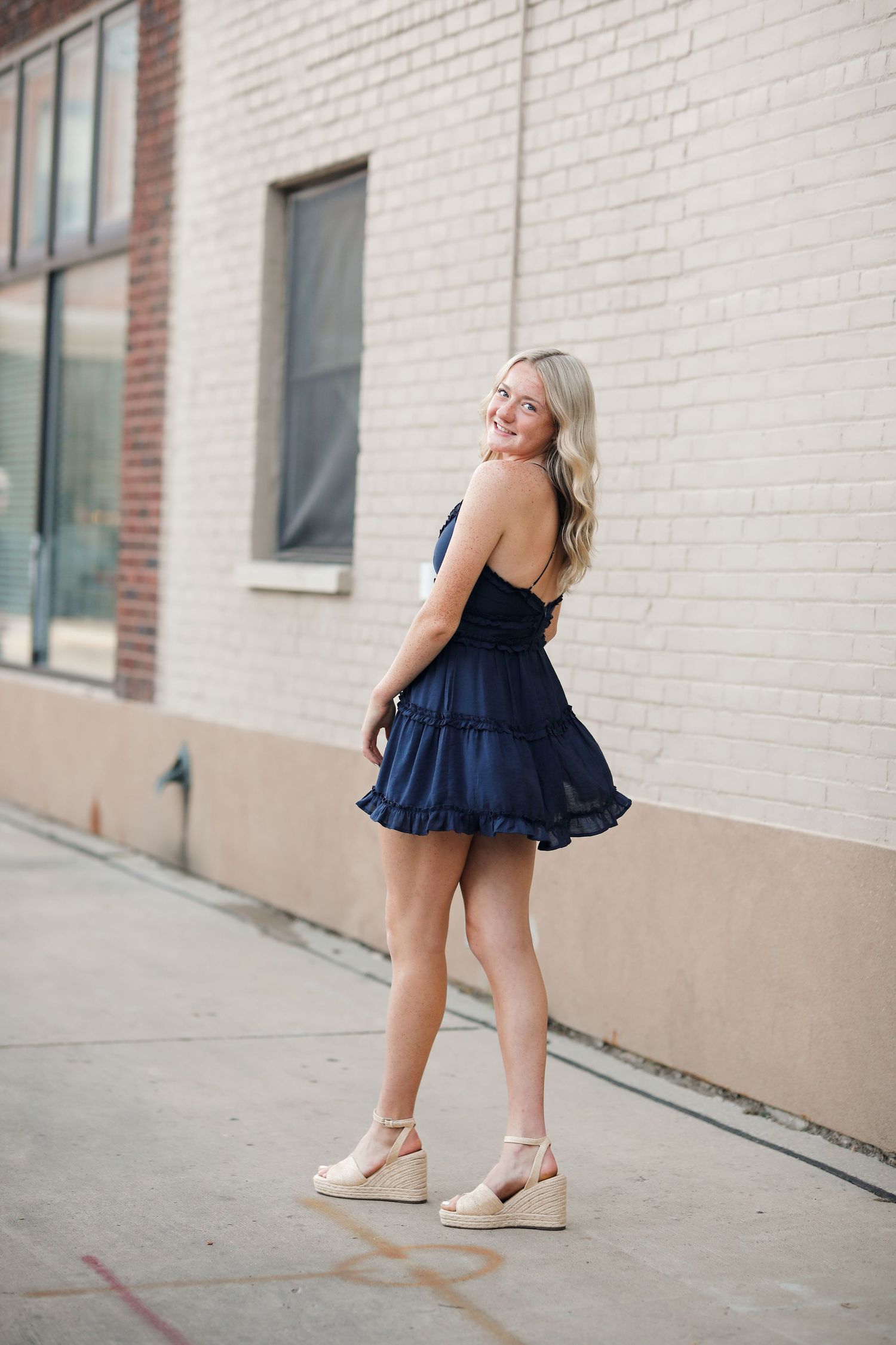 Blonde woman in navy ruffled mini dress and wedge sandals posing on city sidewalk.