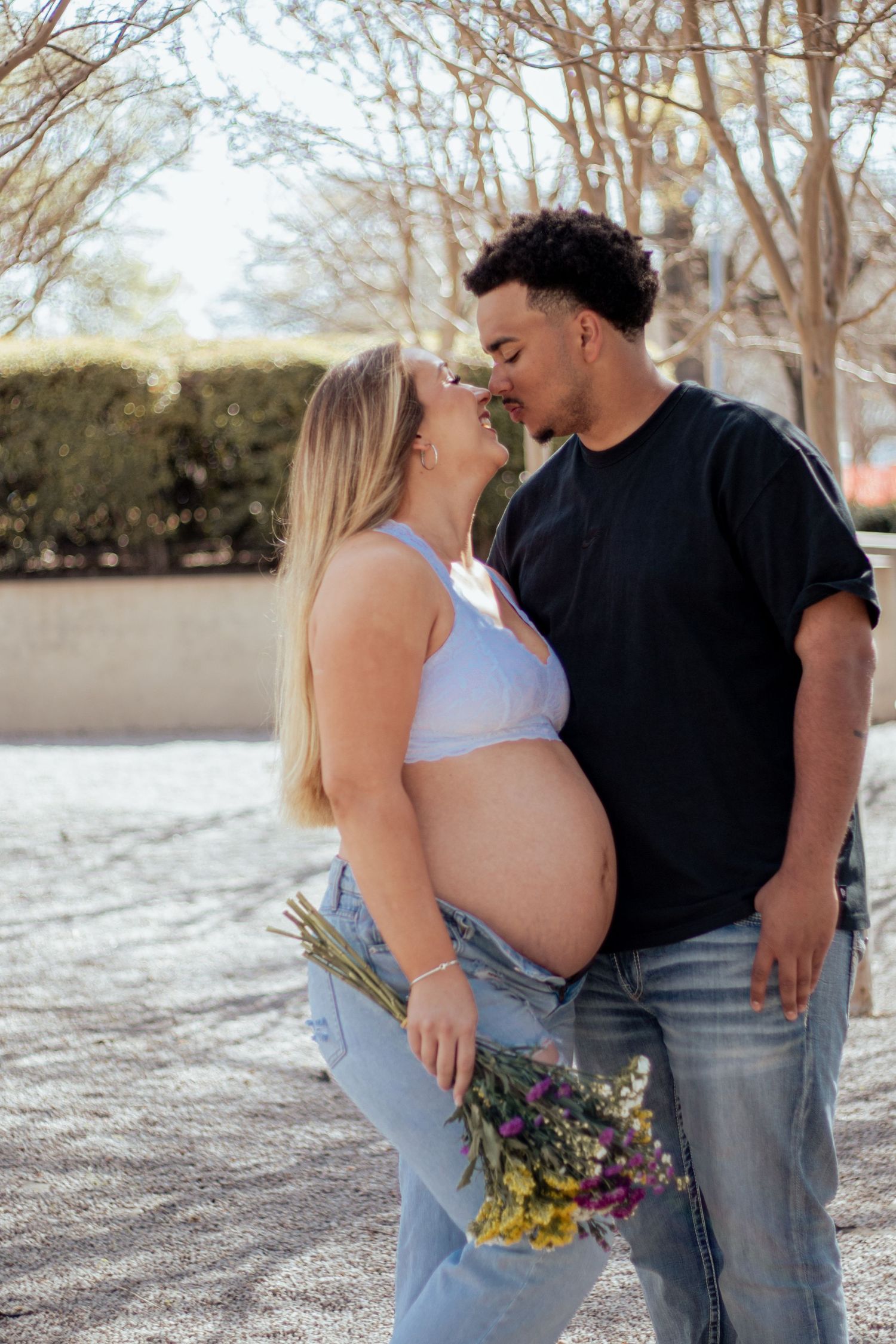 A tender maternity photo outdoors showing an expecting couple sharing an intimate moment with flowers and trees in background.