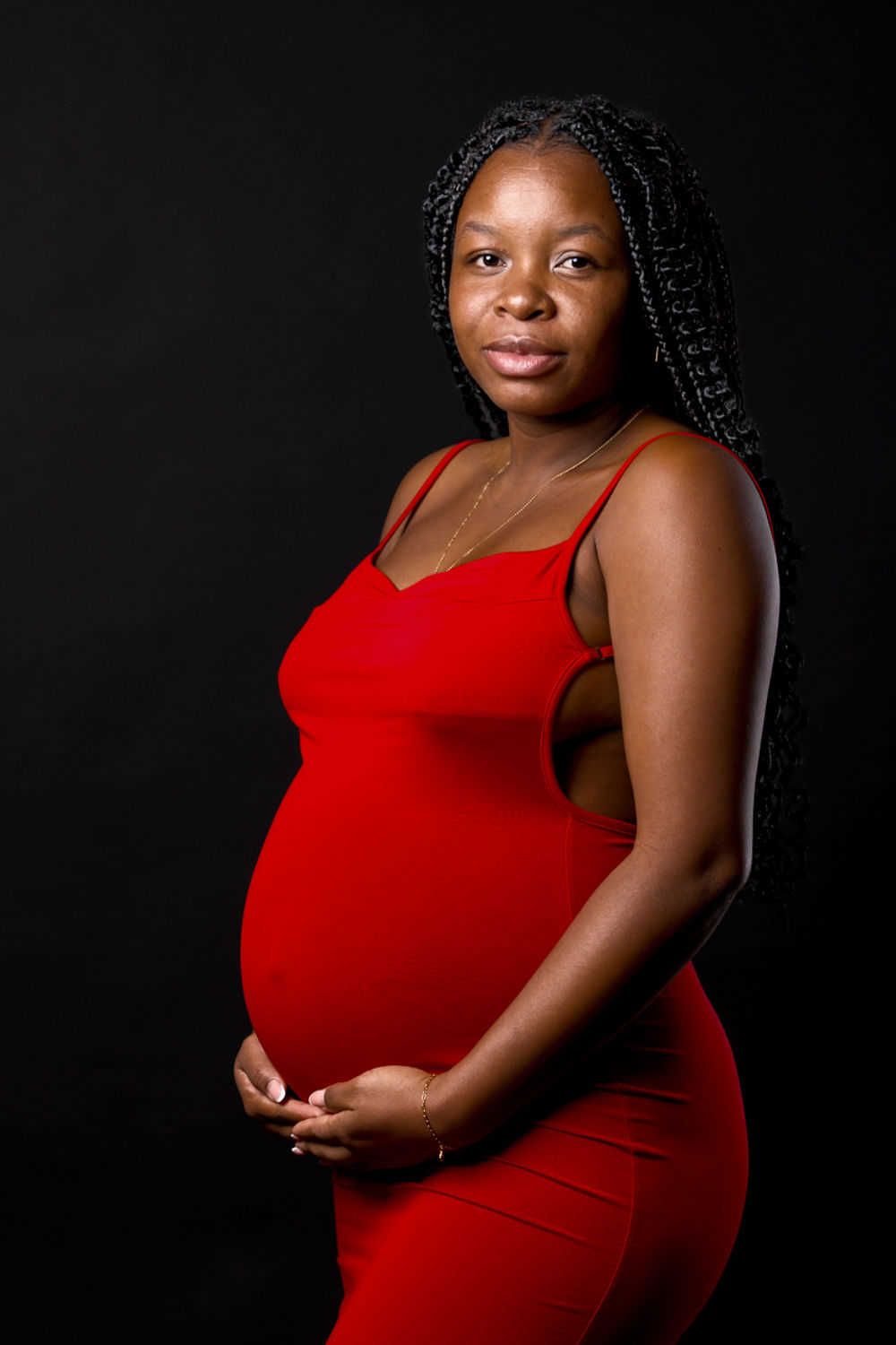 Une femme enceinte vêtue d'une robe rouge ajustée pose avec assurance sur un fond sombre lors d'une séance photo de maternité.