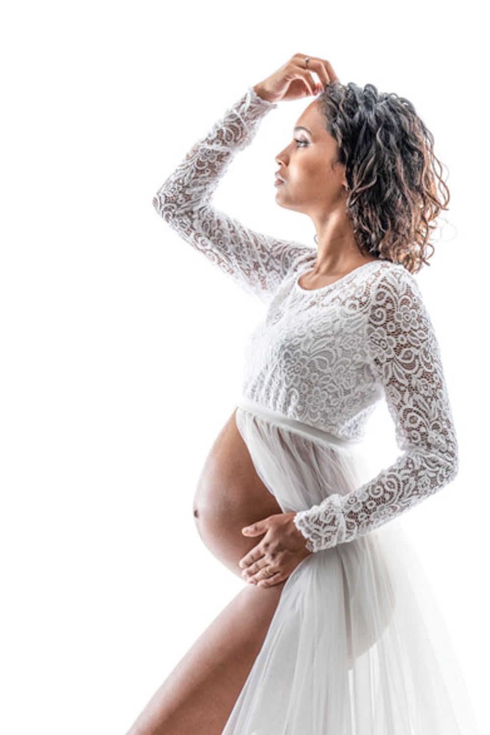 Photo de maternité artistique d'une future maman dans une robe fluide en dentelle blanche posant gracieusement sur un fond clair.