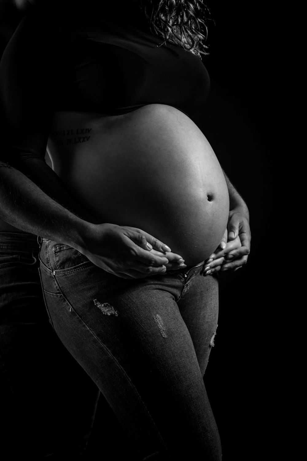 Photo de maternité artistique en noir et blanc montrant un ventre de femme enceinte avec des mains berçant doucement la bosse sur un fond s