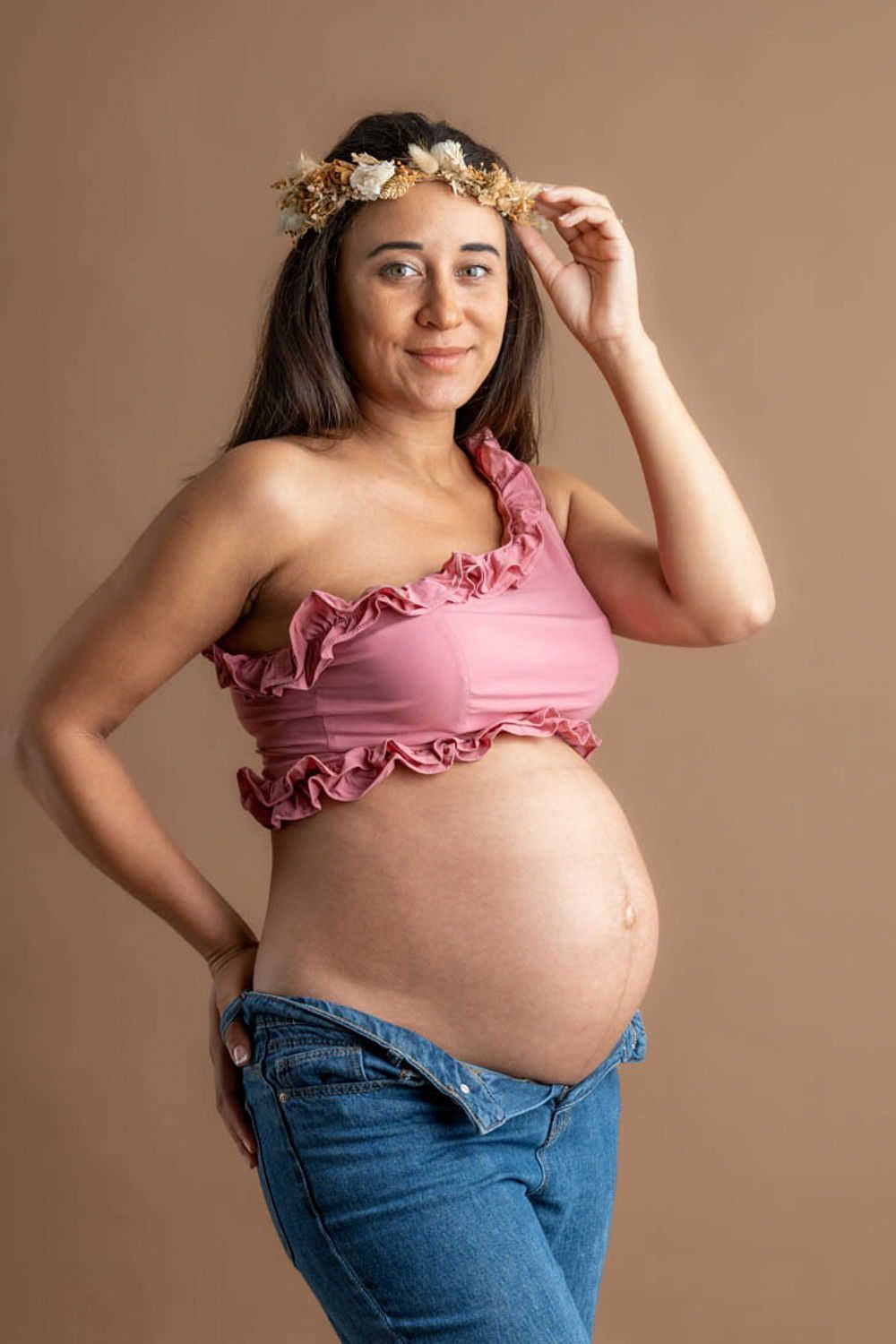 Une future maman portant un haut court à volants rose, une couronne de fleurs et un jean pose sur un fond neutre lors d'une séance photo de