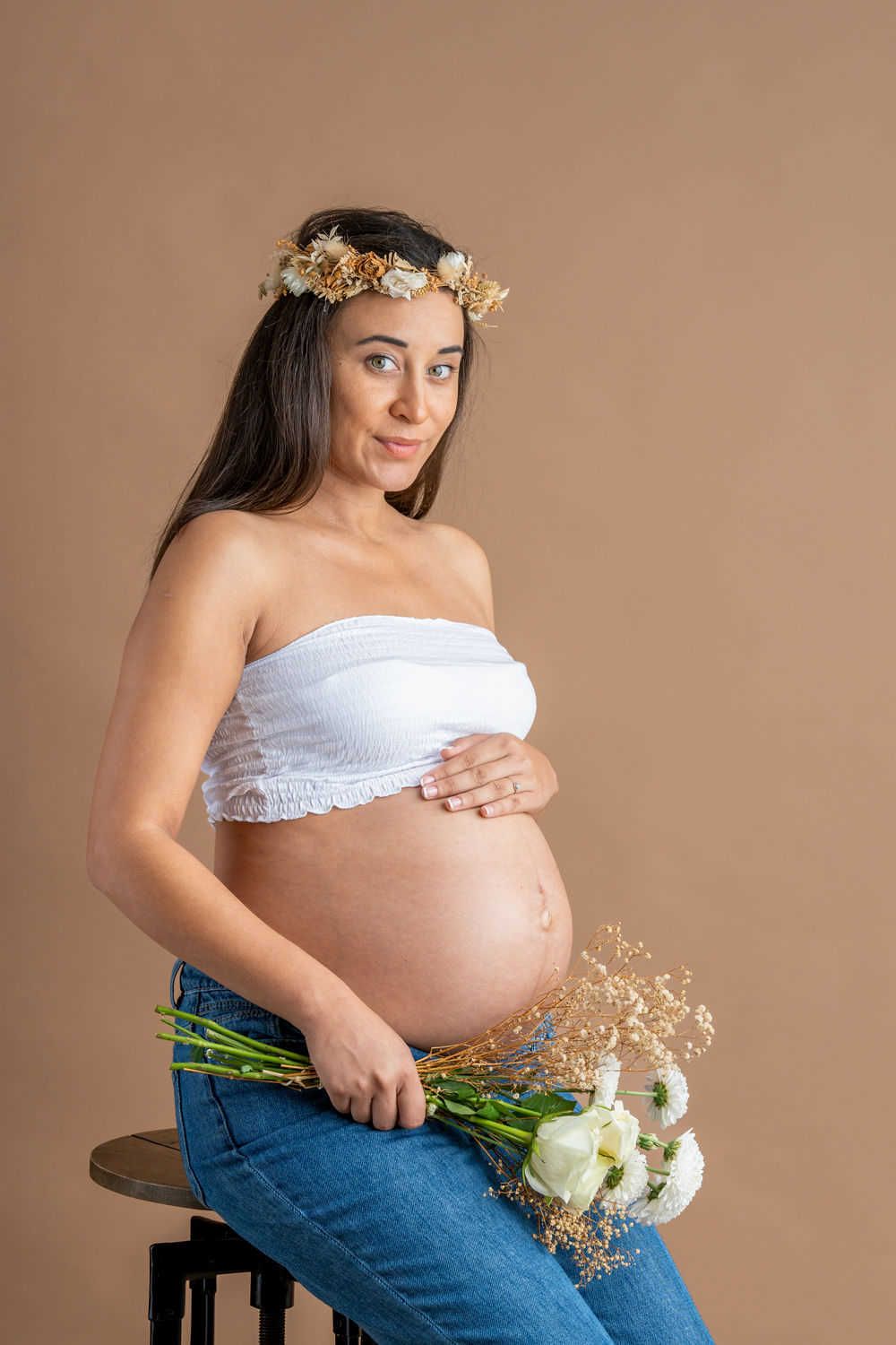 Une future maman en haut bandeau blanc et jean est assise sur un tabouret tenant des fleurs séchées et portant une couronne de fleurs.