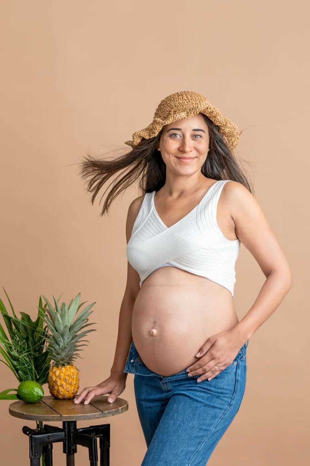 Une femme enceinte portant un chapeau de paille et un jean pose avec des fruits tropicaux sur une table d'appoint sur fond pêche.