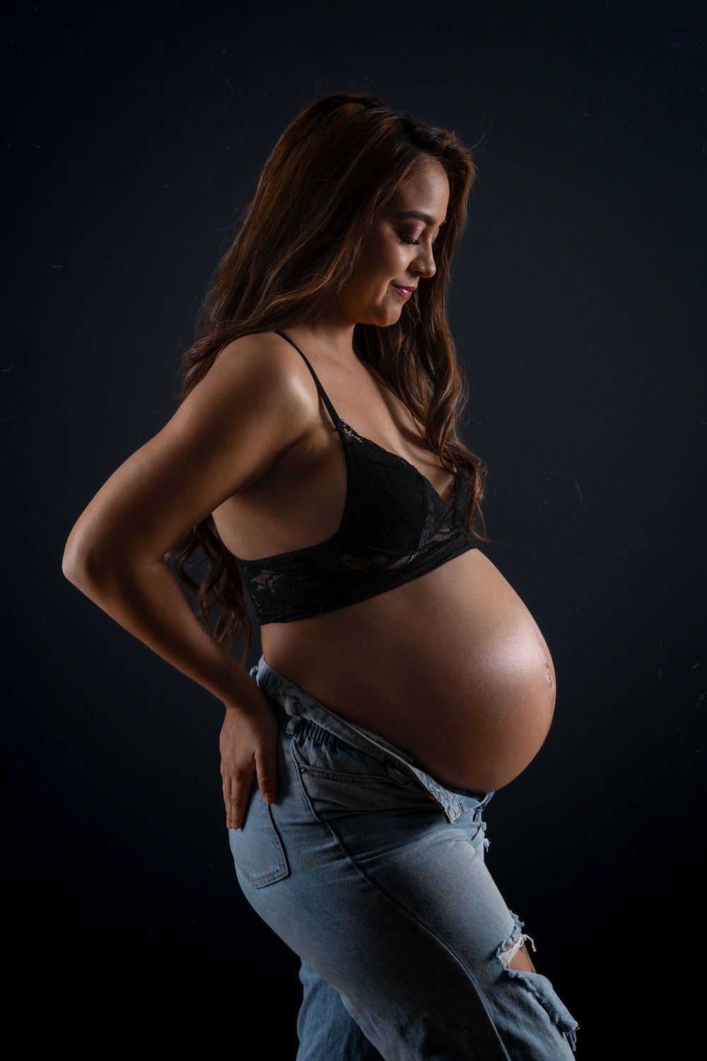 Portrait de maternité vu de côté dans un éclairage tamisé montrant une silhouette enceinte en jean et haut noir sur fond sombre.