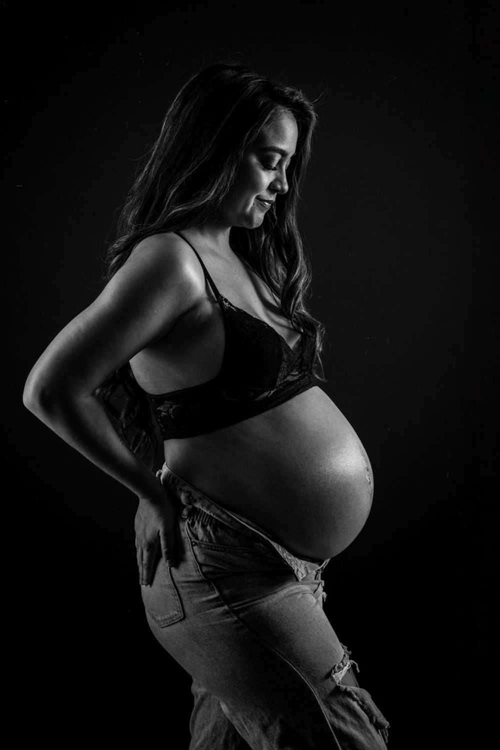Photographie de maternité artistique dramatique en noir et blanc montrant une silhouette enceinte sur un fond sombre.