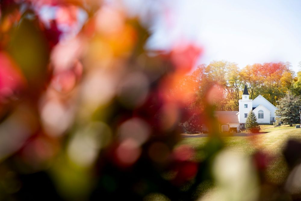 A small white church is softly framed by autumn foliage in the foreground creating a dreamy pastoral scene.   Iowa City wedding photographer