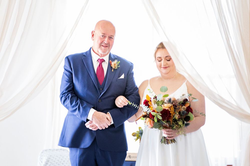 Two people stand together at a wedding ceremony with white curtains, the bride holding a colorful fall bouquet. Iowa City wedding photograph