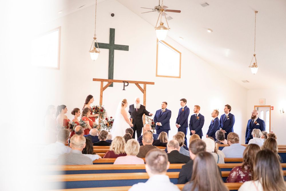 Iowa City wedding photographer. A wedding ceremony takes place in a bright chapel with a wooden cross and rows of guests seated on benches.