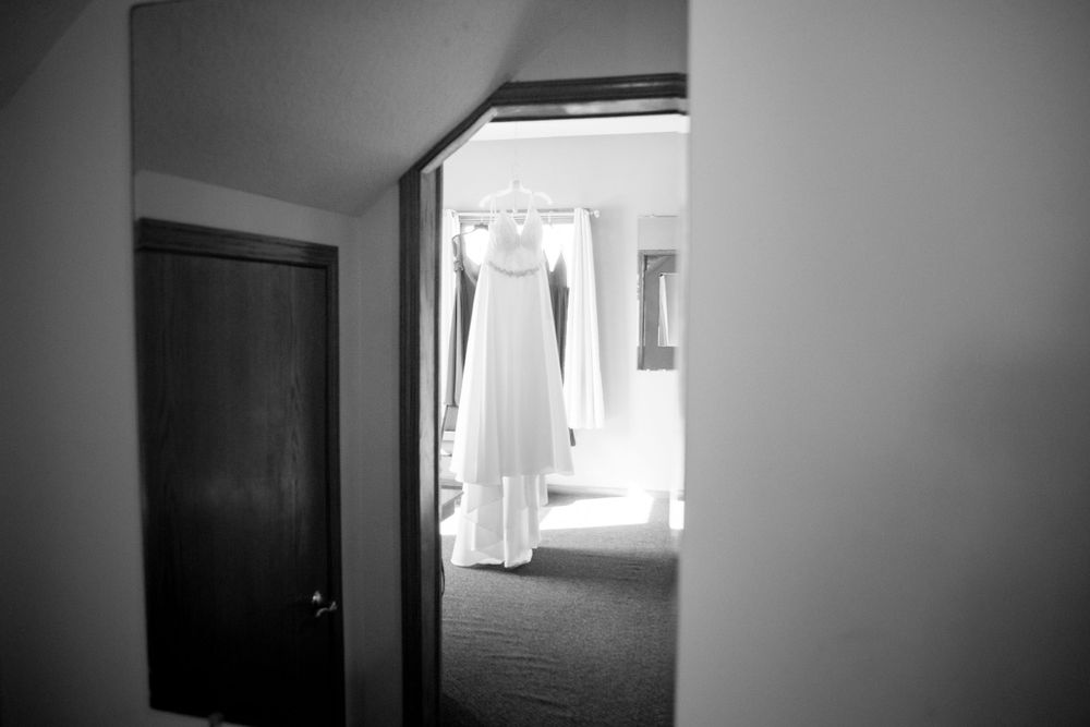 A wedding dress hangs in a doorway, captured  with soft natural lighting creating an ethereal atmosphere. Iowa City wedding photographer.