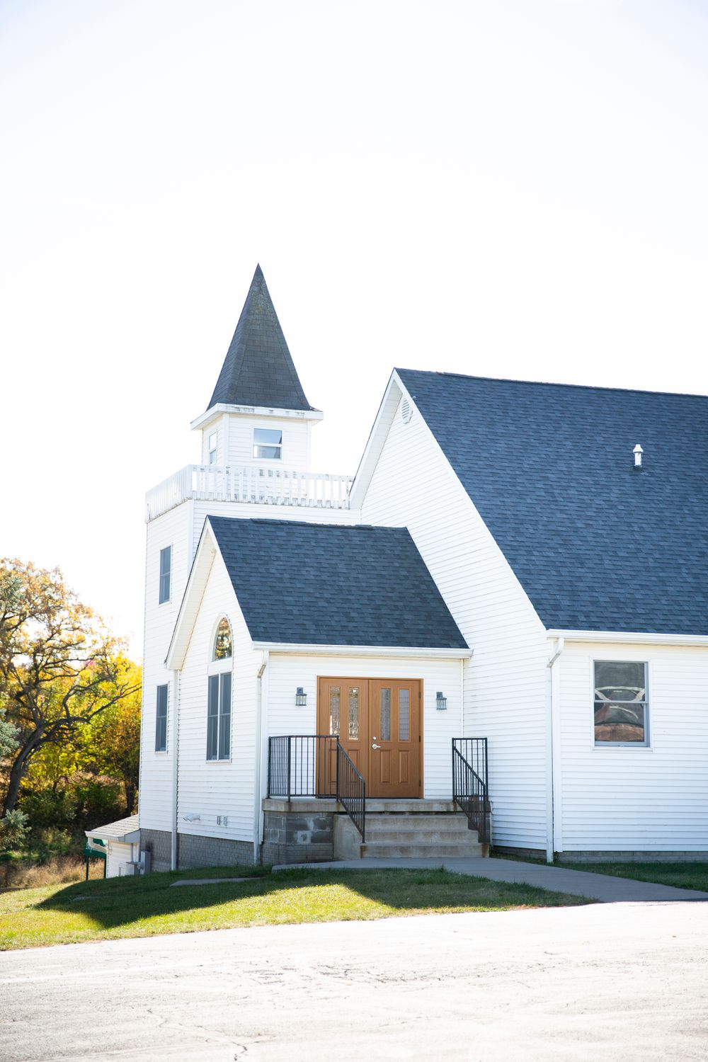 A white country church with a pointed steeple stands against a bright sky on a grassy hillside.  Iowa City wedding photographer.
