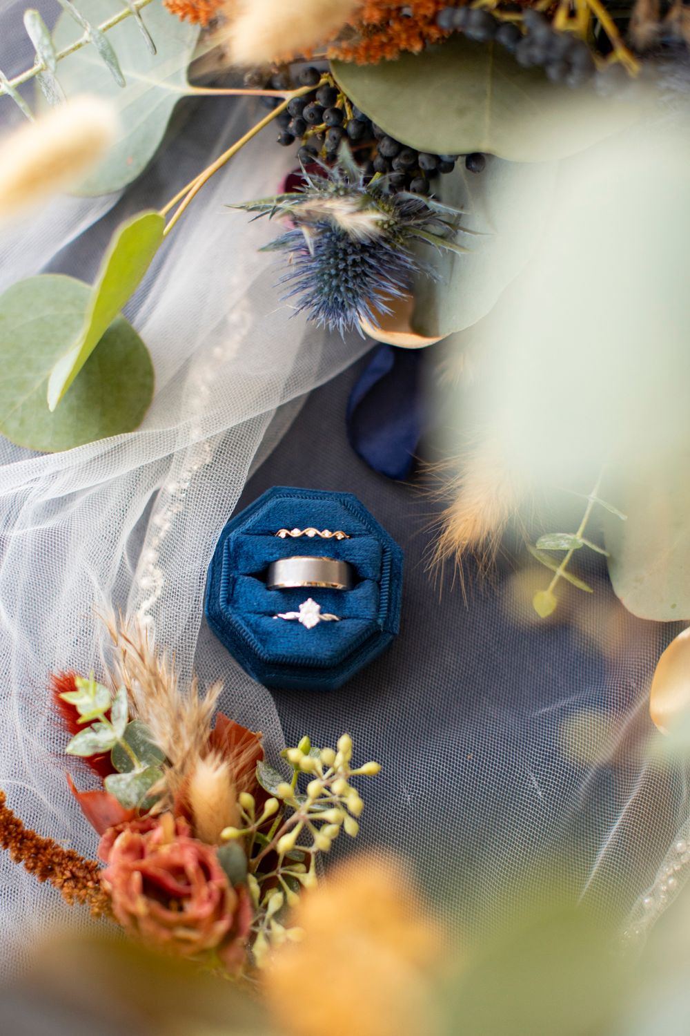 A blue velvet ring box surrounded by delicate floral arrangements and tulle fabric in soft lighting.  Iowa City wedding photographer.