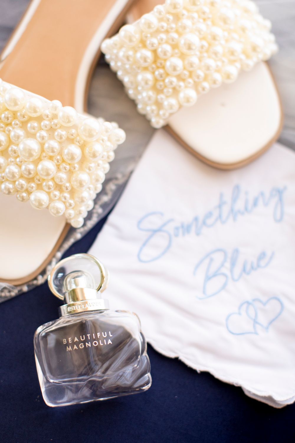 Elegant bridal shoes with pearl details alongside a perfume bottle and Something Blue wedding handkerchief on navy fabric.