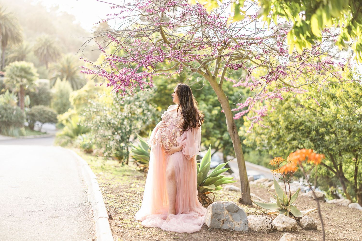 A mom in a flowing pink dress stands beneath blooming cherry blossom trees along a garden path on a sunny day.