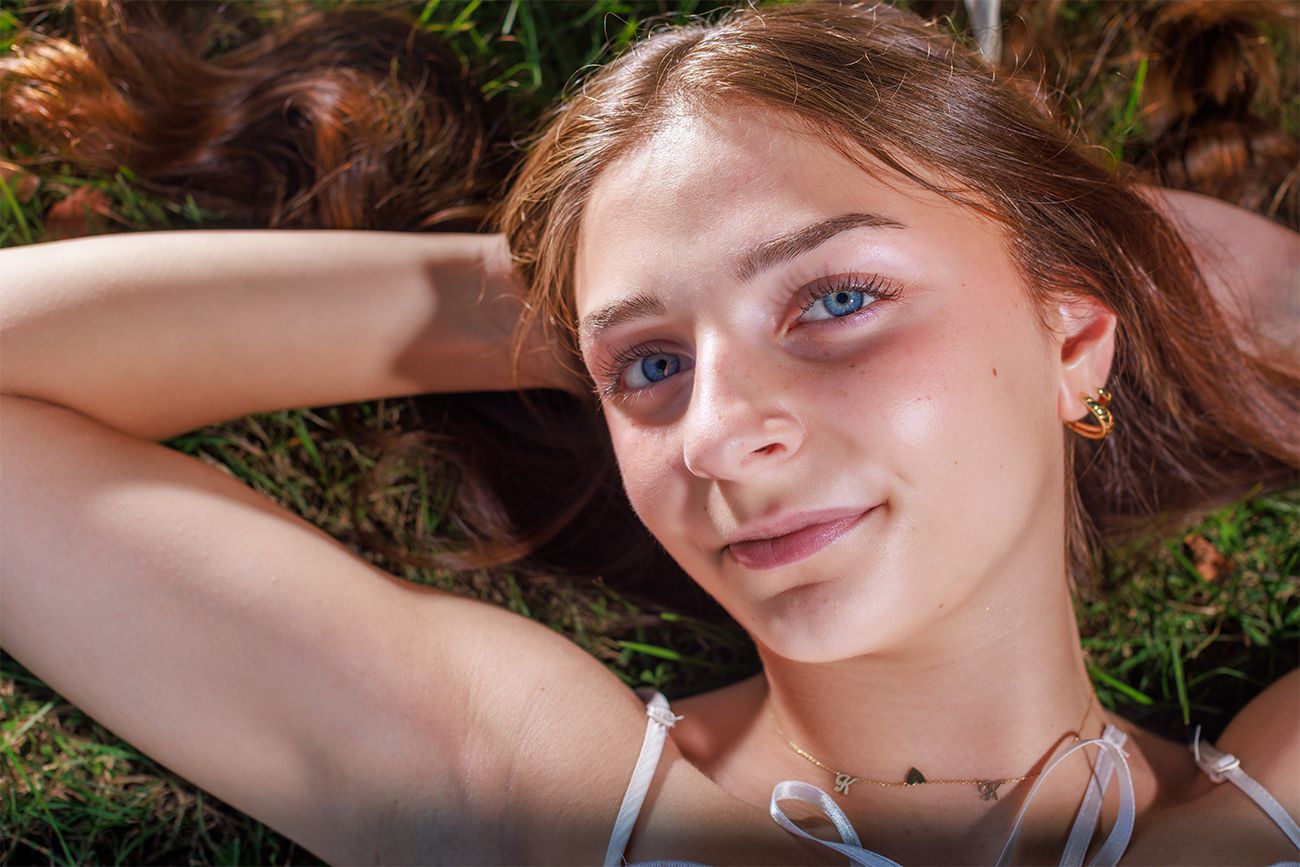 A young person lying in grass smiles at the camera while wearing a white top.