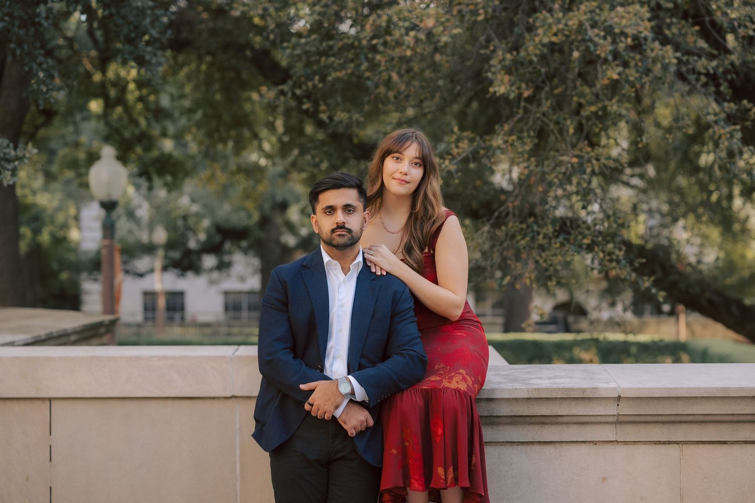 A couple poses together outdoors by a stone wall, with the woman in a red dress standing behind the man in a navy suit.