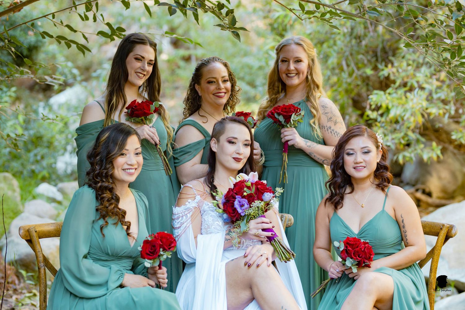 Wedding party in matching seafoam green dresses pose with bright red bouquets in an outdoor garden setting.