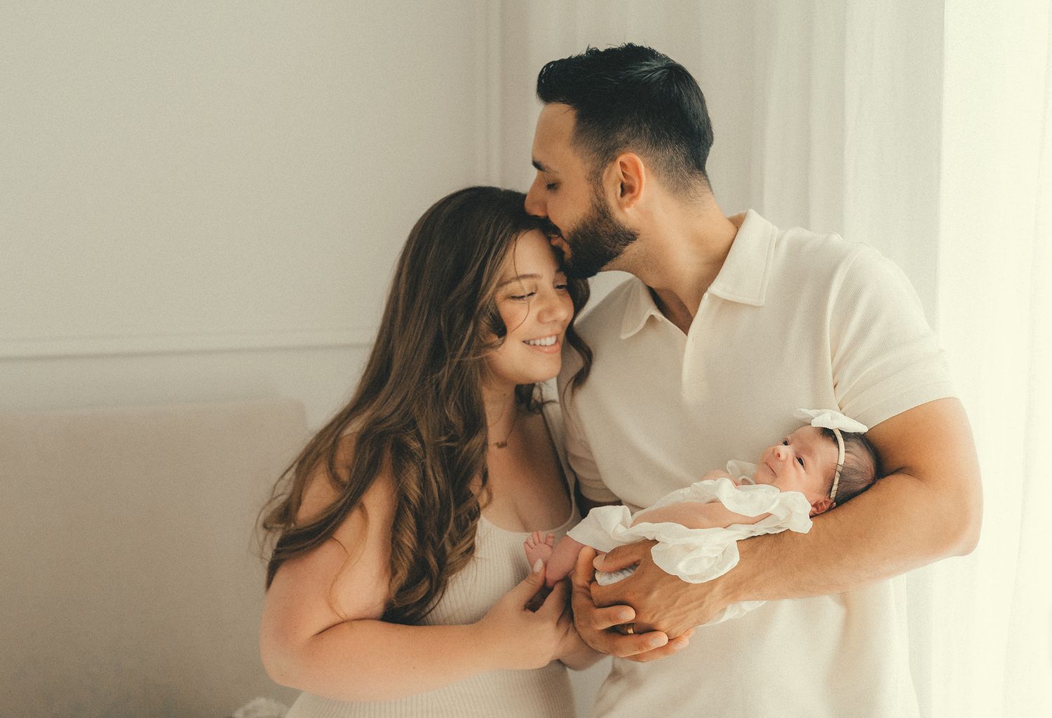 A couple sharing a tender moment while holding their newborn baby in a bright, sunlit room.
