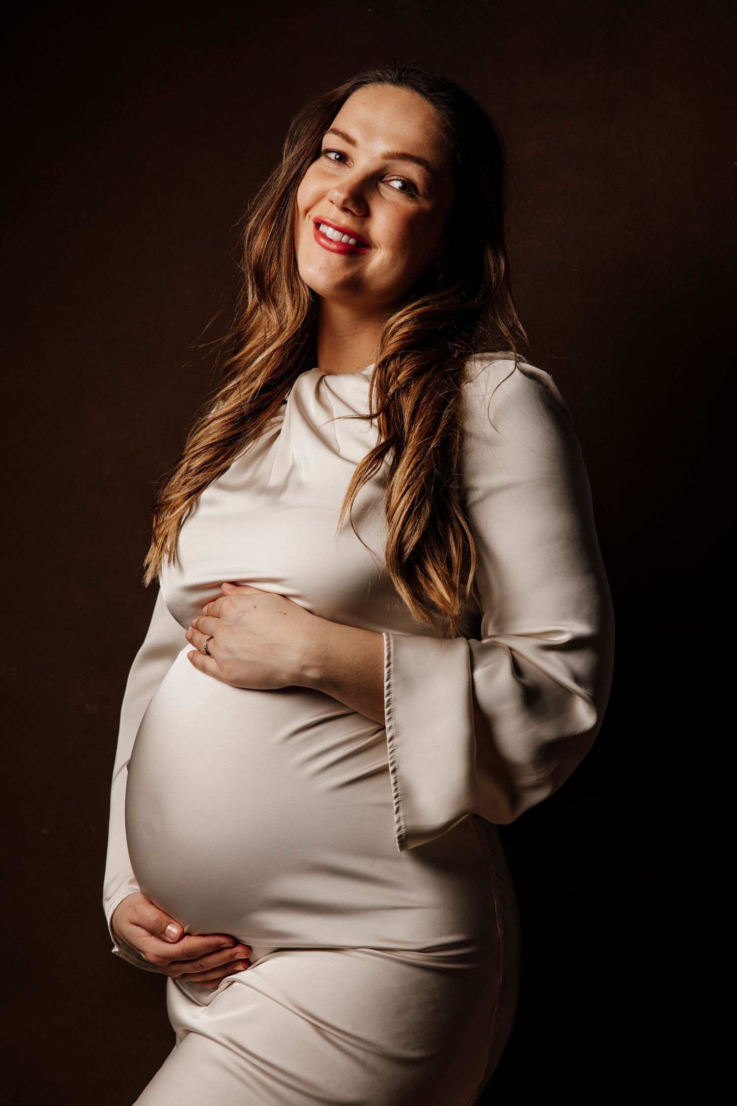 Elegant maternity photo of woman in cream dress against dark background.