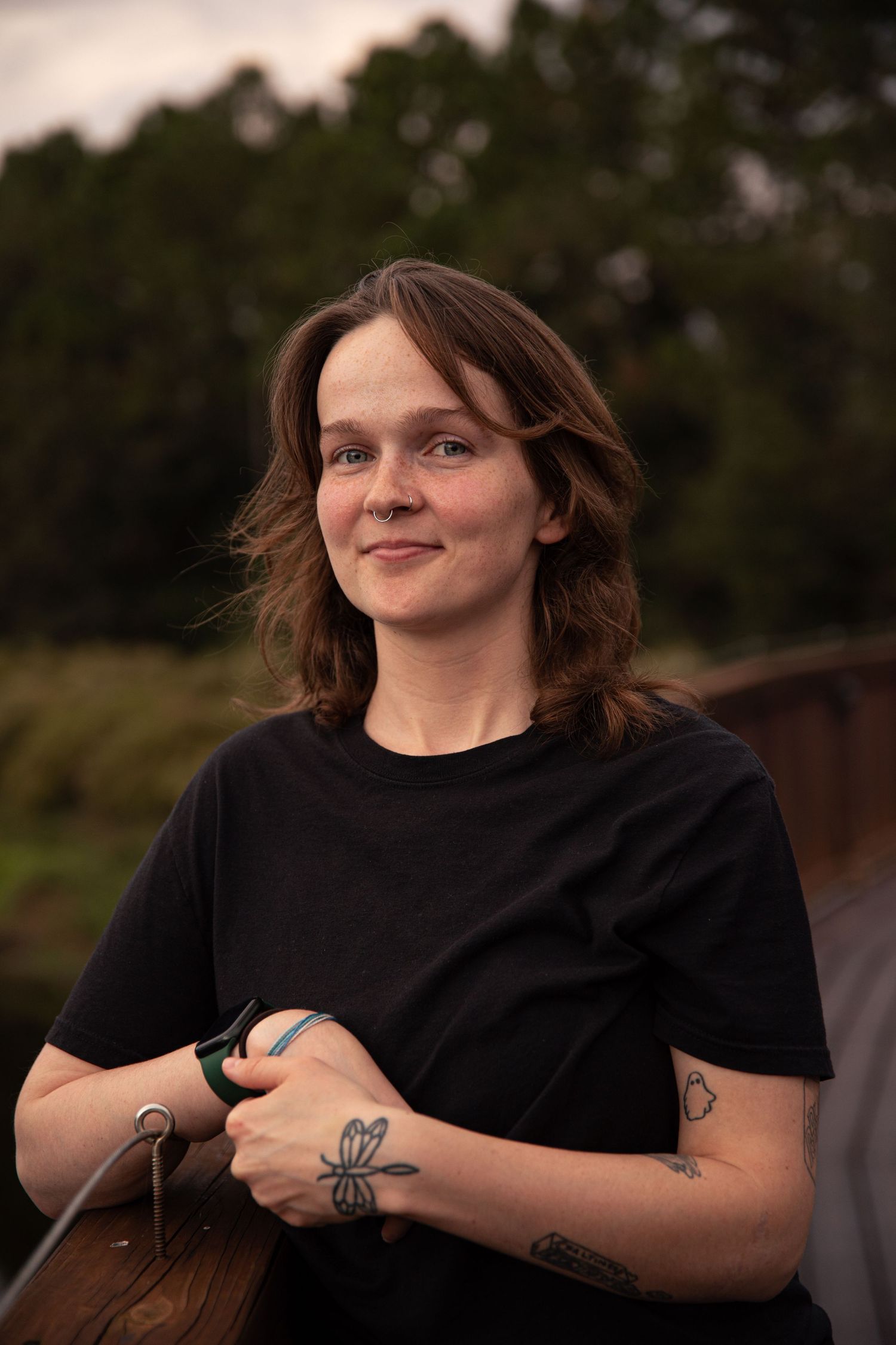 Person in black t-shirt with visible tattoos stands outdoors at dusk.