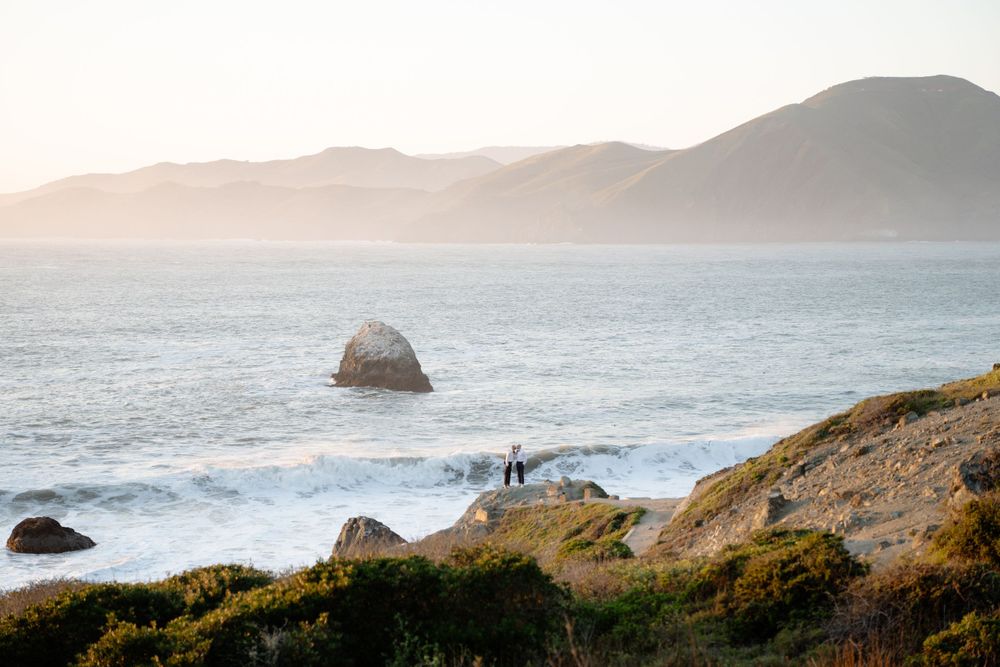 A scenic coastal view shows two people standing on rocky cliffs overlooking the ocean with misty mountains in the distance.